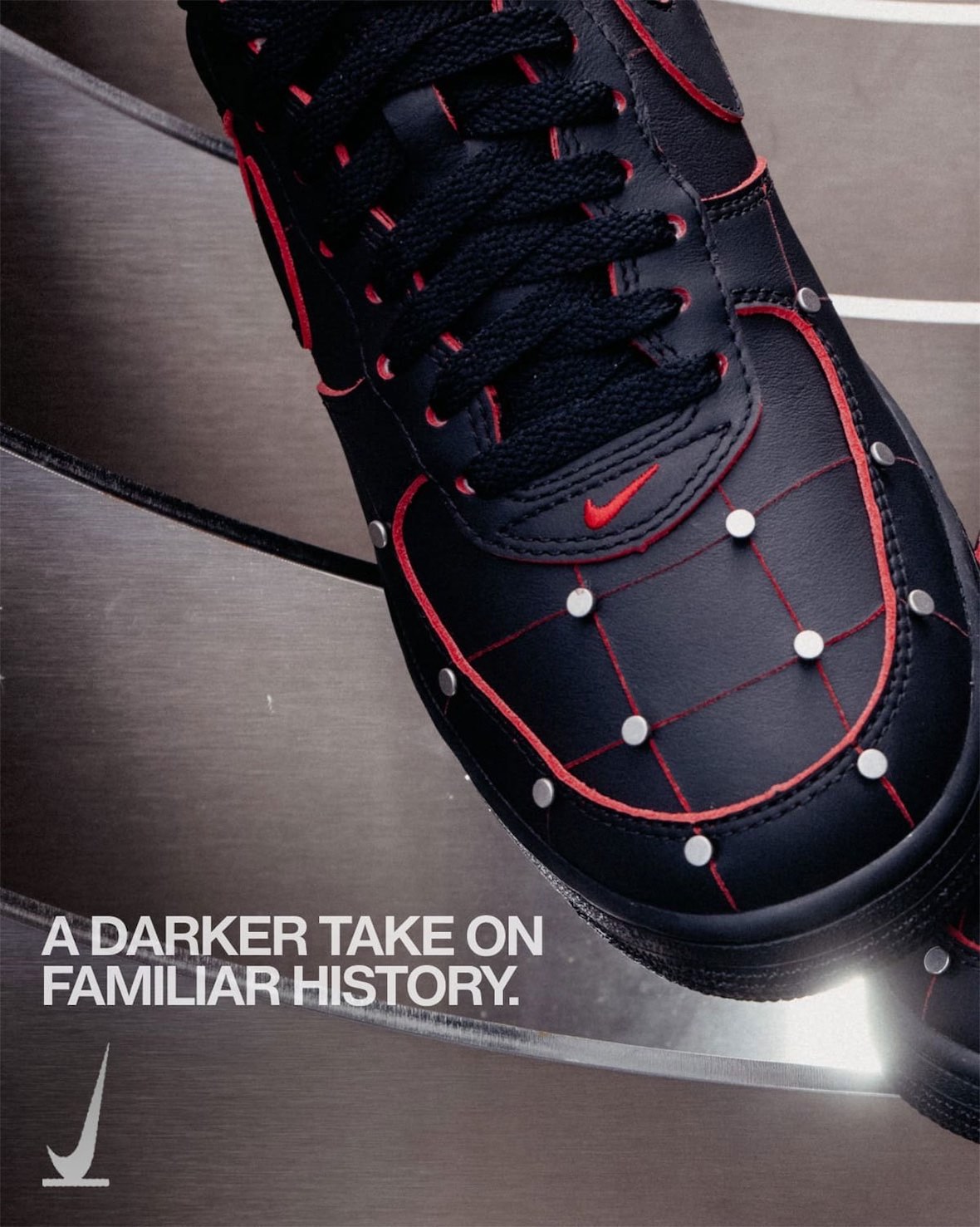 Primer plano de la puntera del Nike Air Force 1 '01 Nightmare en cuero negro, con costuras rojas que forman una cuadrícula, tachas plateadas en cada cruce, cordones negros, ojales con borde rojo y swoosh rojo chico, sobre una superficie metálica curva