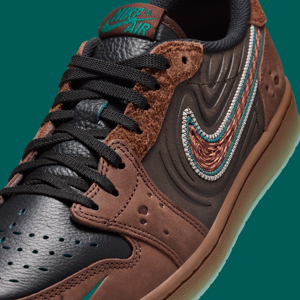 Primer plano del lateral del Air Jordan 1 Low OG Somos Historias, con los cordones negros, la gamuza marrón de pelo largo en los ojales, el cuero marrón oscuro con relieves en ondas, el swoosh tejido marrón con borde verde agua y gris, y el logo Nike Air verde en la lengüeta
