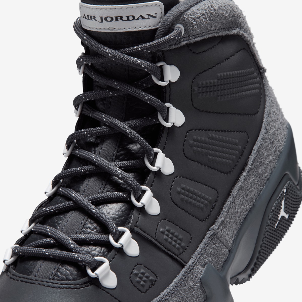 Primer plano del Air Jordan 9 Retro Boot NRG Anthracite mostrando los cordones gris oscuro con puntos claros pasando por los ojales metálicos en forma de D con terminales grises, la etiqueta Air Jordan en la lengüeta, el cuero negro con relieves y la gamuza gris de pelo largo