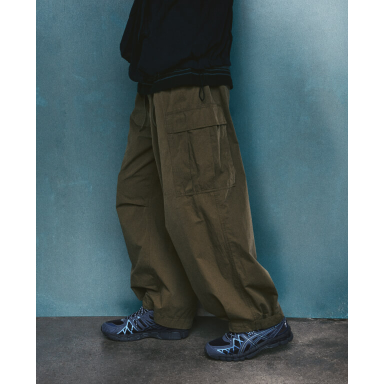 Foto de campaña de la ASICS Utility Collection con el GEL-KAYANO 20 en azul marino oscuro con la jaula de TPU en celeste, usado con un pantalón cargo verde oliva frente a una pared azul