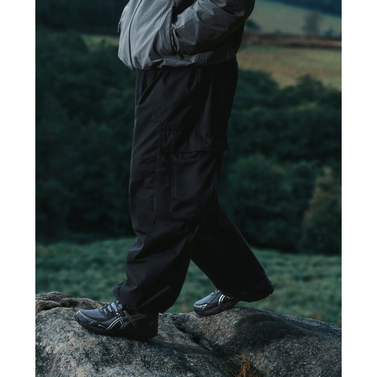 Foto de campaña de la ASICS Utility Collection con el GEL-VENTX GTX gris y negro con las rayas ASICS plateadas, usado con pantalón cargo negro sobre una roca en un paisaje de campo