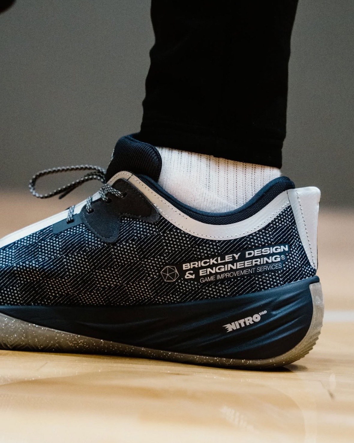 Costado de la PUMA All-Pro Nitro 2 Race 2 Evolve de Chris Brickley puesta en una cancha de madera, con la leyenda Brickley Design & Engineering, el logo Nitro en el talón y la suela gris con puntitos