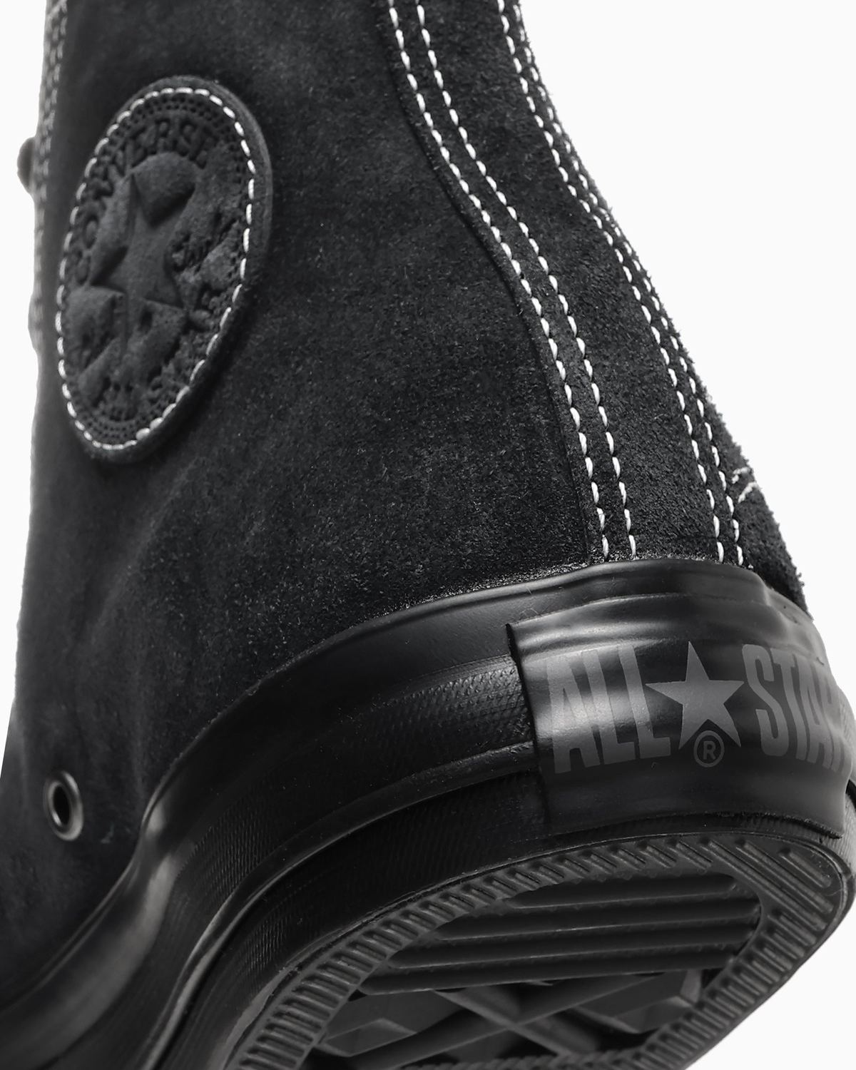 Primer plano del talón de la versión negra: gamuza negra con pespuntes blancos, el parche circular del tobillo en relieve y la etiqueta de goma del talón con el logo ALL STAR en gris sobre la suela negra