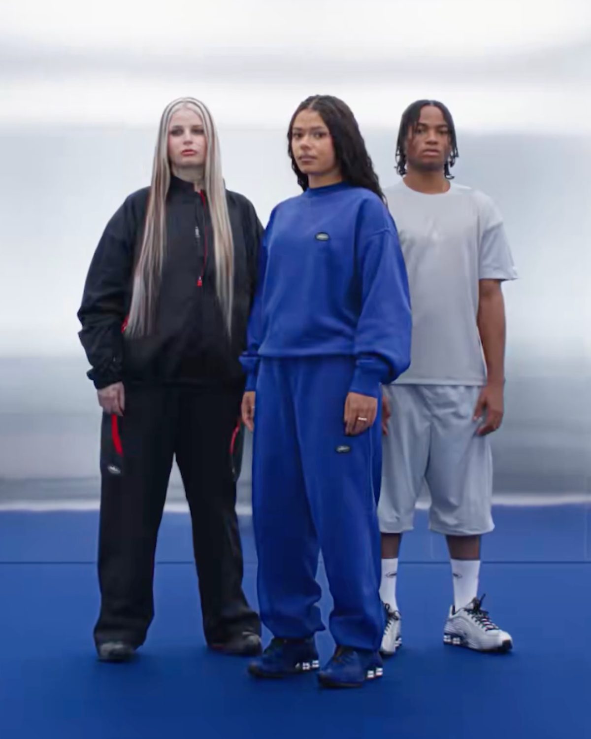 Tres modelos de la campaña de Corteiz x Nike de pie sobre un piso azul: una con conjunto negro, otra con conjunto azul y un chico con remera gris y shorts, cada uno con un color distinto del Shox R419