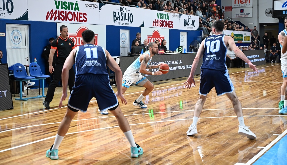 Un jugador de Racing de Chivilcoy, con camiseta blanca y celeste, se prepara para pasar la pelota ante la marca de dos jugadores de Oberá con camisetas azules, en la cancha del Oberá Tenis Club