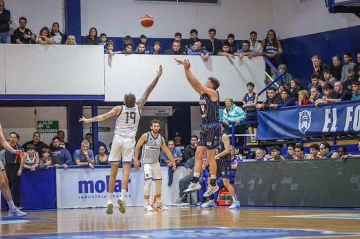 Un jugador de Argentino de Junín, de azul oscuro, lanza en suspensión ante la marca de un jugador de Boca, de blanco, con la tribuna local de fondo