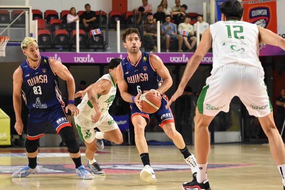 Un jugador de Quimsa con la camiseta azul 6 maneja la pelota ante la marca de jugadores de Atenas con camiseta blanca y verde, en el Estadio Ciudad de Santiago del Estero