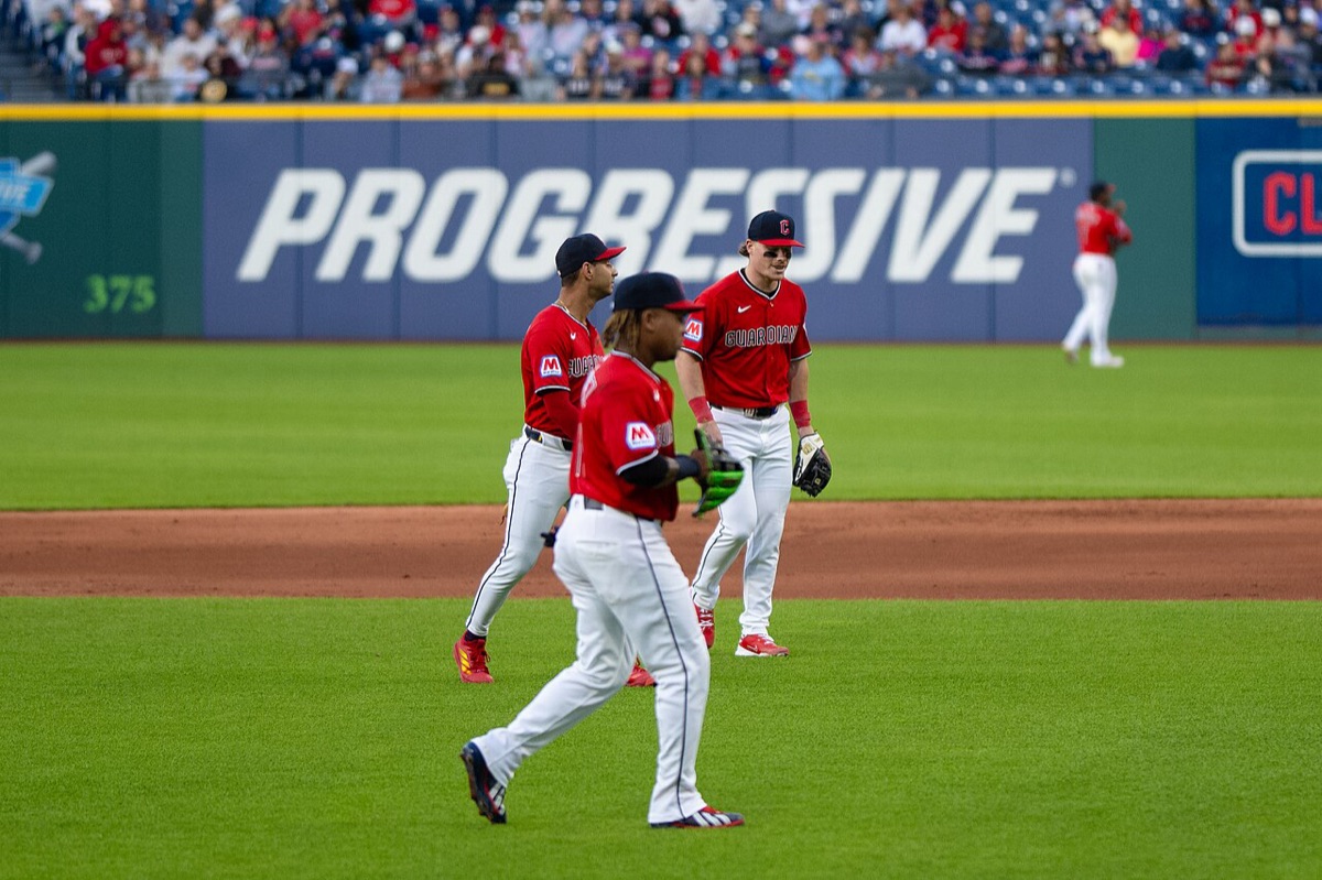 Foto de archivo: tres jugadores de los Cleveland Guardians, con camiseta roja y pantalón blanco, caminan por el césped de Progressive Field, con un cartel azul de Progressive en la pared del outfield