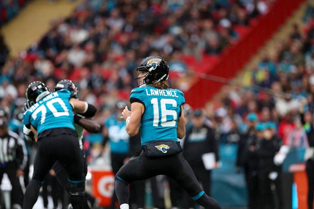 Trevor Lawrence, con la camiseta turquesa número 16 de los Jaguars, prepara un pase detrás de su línea, con el número 70 bloqueando a su izquierda y la tribuna de Wembley detrás