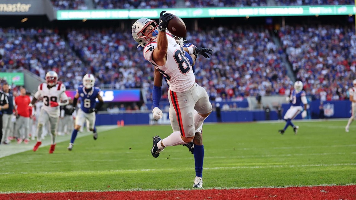 Un receptor de los Patriots, de blanco, estira un brazo para atrapar la pelota frente a un defensor de los Bills cerca de la zona de anotación del Highmark Stadium