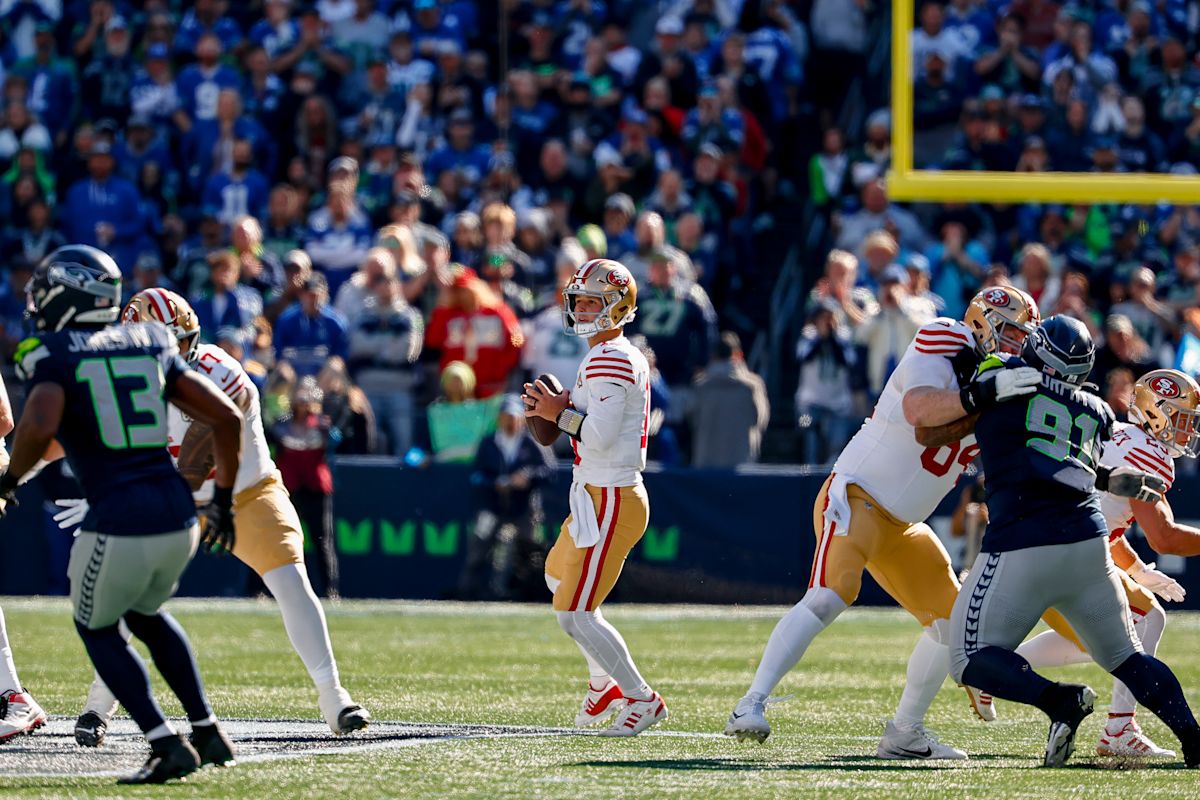 Brock Purdy, quarterback de los 49ers, con camiseta blanca, se prepara para lanzar en el Lumen Field mientras su línea lo protege de los Seahawks