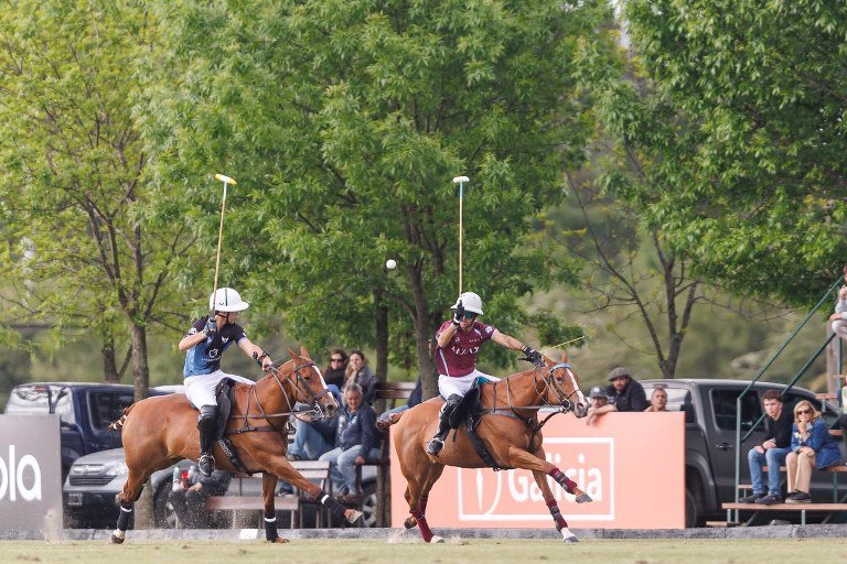 Dos jugadores, uno de camiseta azul y otro bordó, galopan hacia la bocha frente a una tribuna con público en una cancha de polo