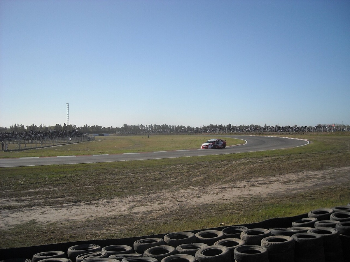 Foto de archivo: un auto de carrera rojo y blanco dobla una curva del autódromo de Rosario, con público en las tribunas del fondo y una barrera de cubiertas adelante
