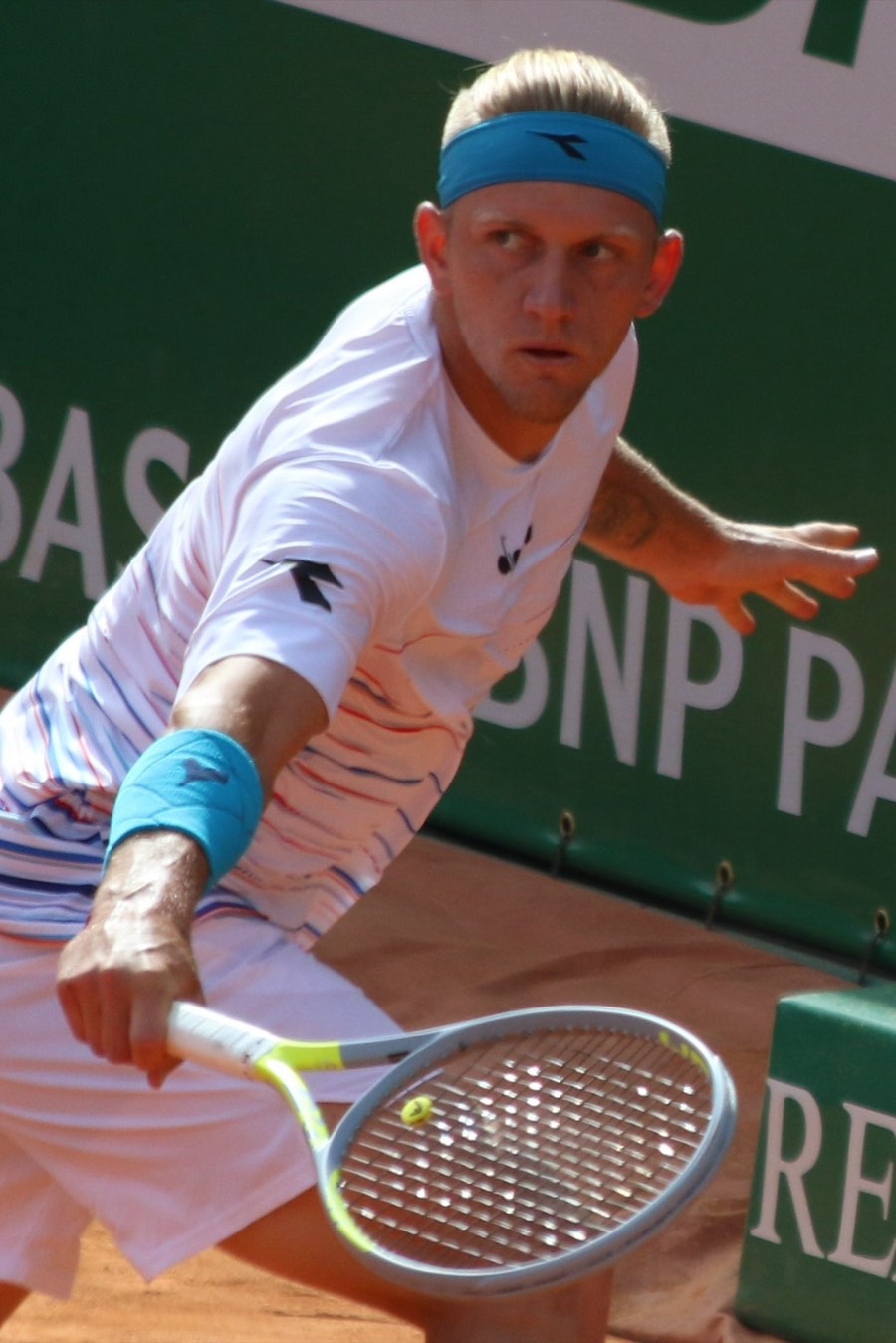 Foto de archivo: Alejandro Davidovich Fokina, con vincha celeste y remera blanca a rayas, le pega un revés en una cancha de polvo de ladrillo, con un cartel verde detrás