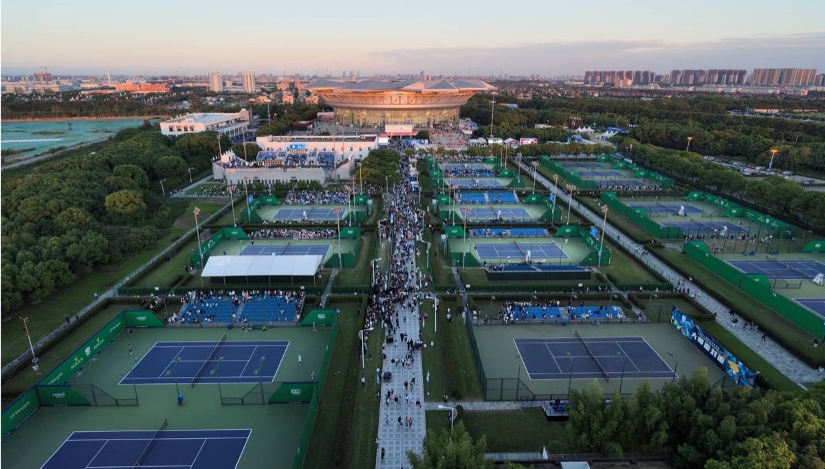 Vista aérea del Qi Zhong Tennis Center de Shanghái al atardecer, con las canchas exteriores azules y verdes, el público en el pasillo central y el estadio principal al fondo