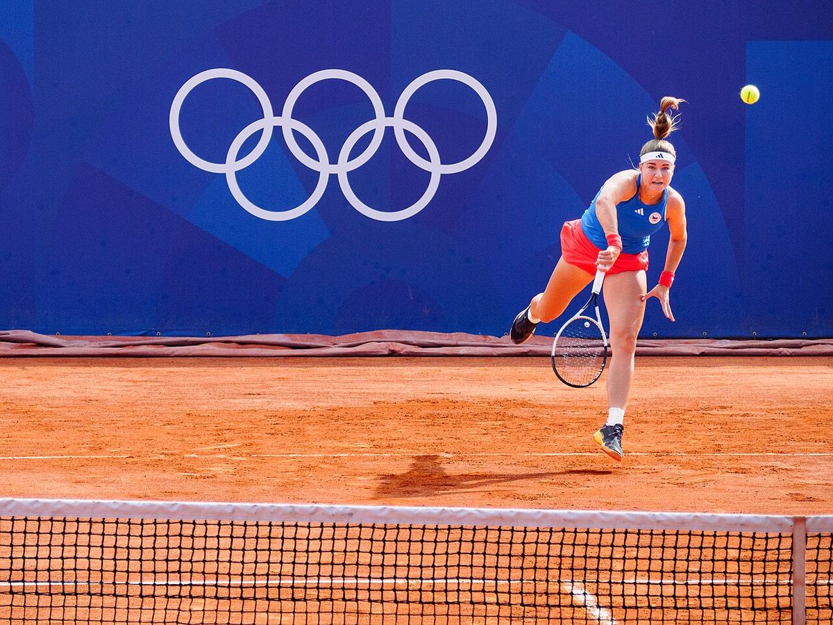 Foto de archivo: Karolína Muchová, con remera azul y pollera roja, saca en una cancha de polvo de ladrillo con los anillos olímpicos de fondo