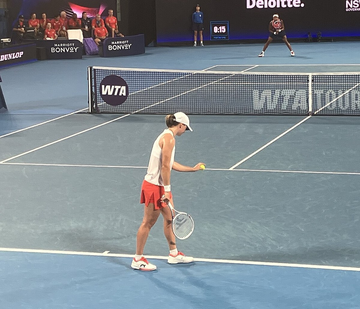 Foto de archivo: Iga Swiatek, con gorra blanca, remera blanca y pollera roja, se prepara para sacar en la semifinal de la United Cup 2026, con Coco Gauff al fondo