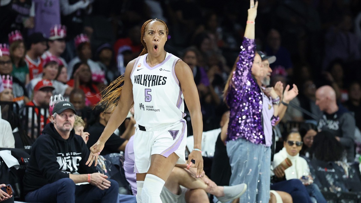 Kayla Thornton, con la camiseta blanca número 5 de Golden State Valkyries, grita y festeja un triple frente a la tribuna, con una hincha vestida de violeta aplaudiendo atrás