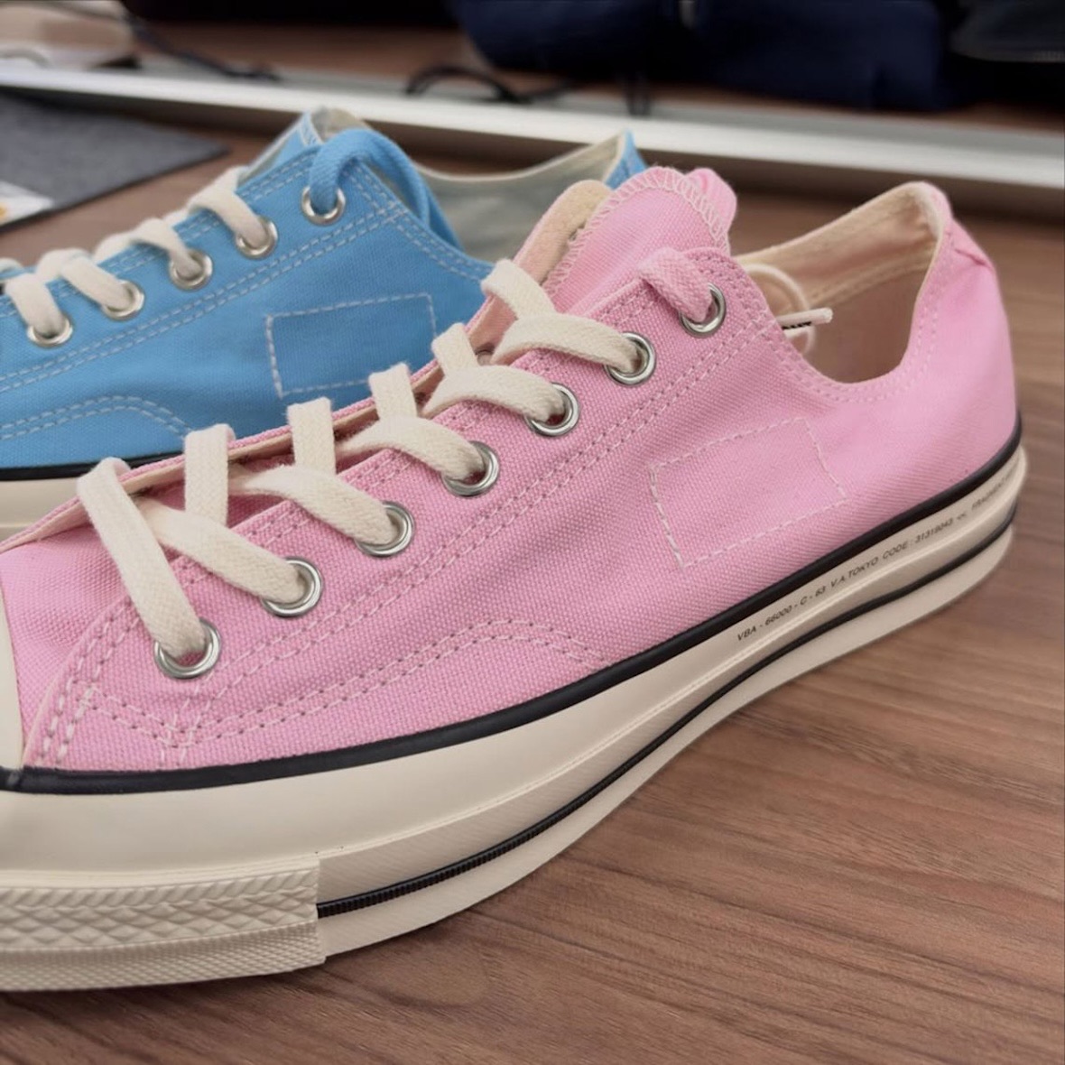 Converse Chuck Taylor baja de fragment design en rosa, de perfil sobre una mesa de madera, con un par celeste detrás y una costura rectangular en la capellada