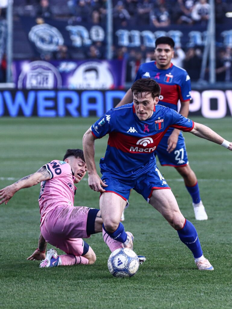 Un jugador de Tigre, de azul y rojo, se lleva la pelota ante un futbolista de Independiente Rivadavia de camiseta rosa que se barre (foto: Liga Profesional de Fútbol)
