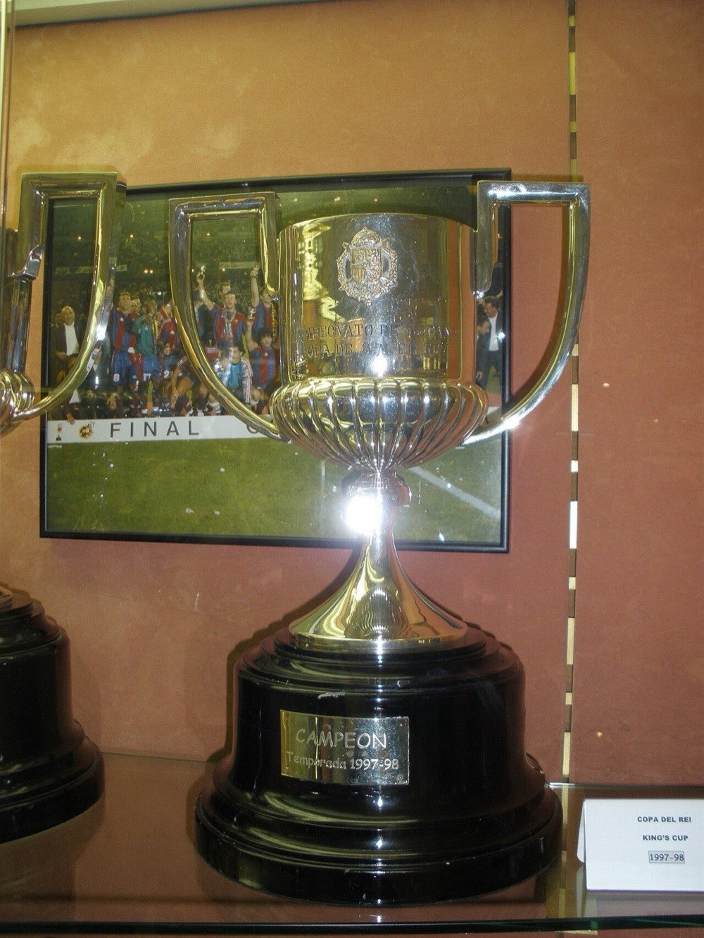 El trofeo de la Copa del Rey, una copa plateada con dos asas sobre una base negra, exhibido en una vitrina con la placa de campeón de la temporada 1997-98 (foto de archivo: Ignasi, Wikimedia Commons, CC BY-SA 4.0)