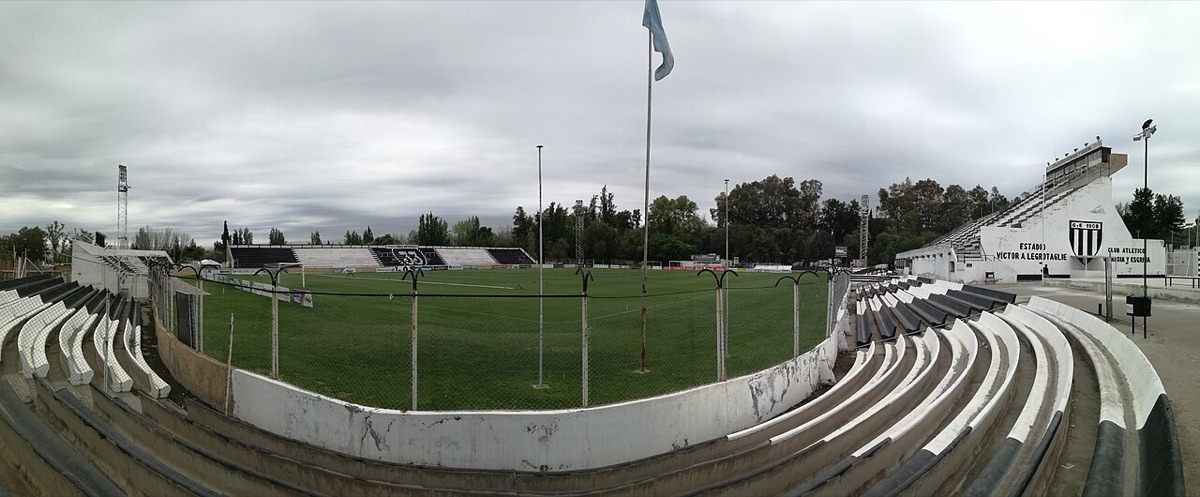 Vista panorámica lateral del estadio Víctor Antonio Legrotaglie, de Gimnasia y Esgrima de Mendoza, con las gradas blancas y negras y el escudo del club sobre la tribuna (foto de archivo de 2017: Iván Aguilar, Wikimedia Commons, CC BY-SA 4.0)