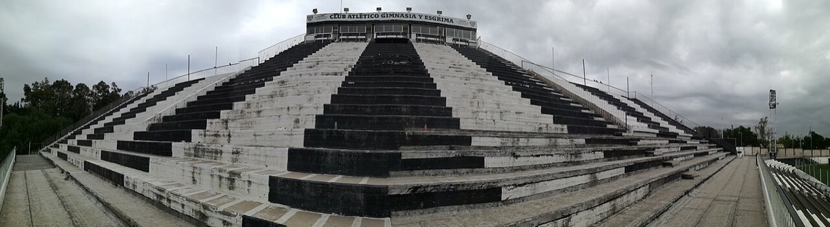 La platea oeste del estadio Víctor Antonio Legrotaglie, de Gimnasia y Esgrima de Mendoza, con sus gradas pintadas de blanco y negro y el nombre del club en lo alto; foto de archivo de 2017 (foto: Iván Aguilar / Wikimedia Commons, CC BY-SA 4.0)