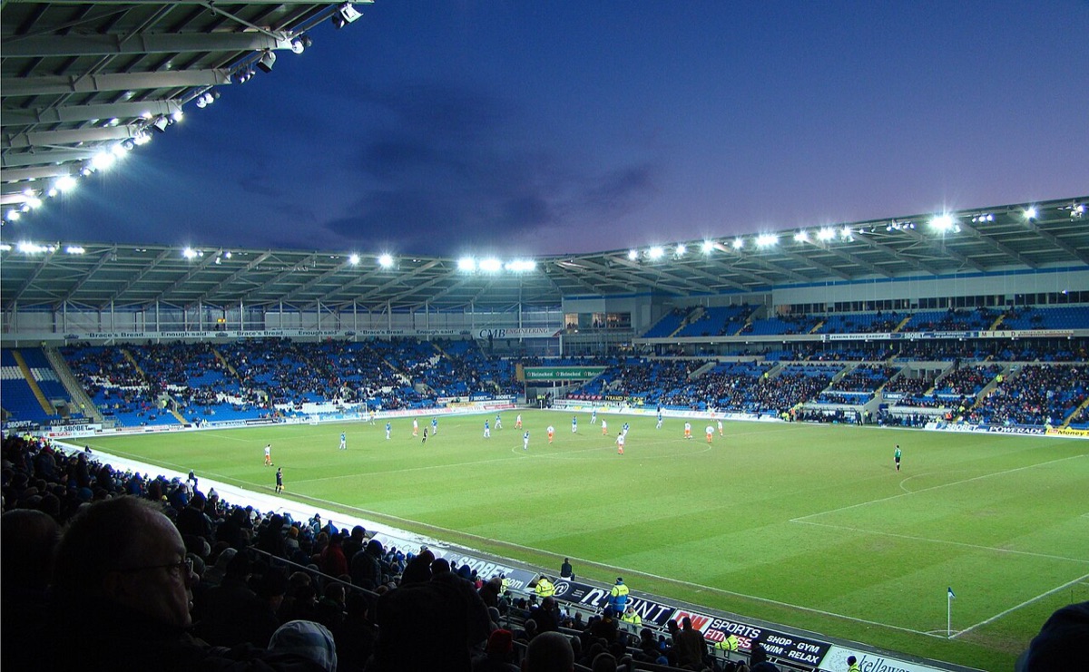 El Cardiff City Stadium iluminado de noche durante un partido, con las tribunas azules y el campo de juego (foto de archivo: Jon Candy, Wikimedia Commons, CC BY-SA 2.0)