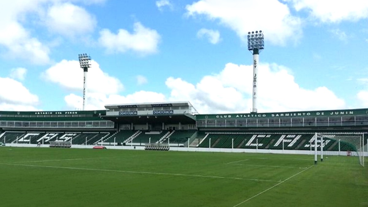 El estadio Eva Perón de Junín visto desde el campo de juego, con la platea verde y blanca de Sarmiento, las letras CAS en las gradas, el arco a la derecha y dos torres de iluminación bajo un cielo con nubes; foto de archivo de 2015 (foto: Paolazaccardi / Wikimedia Commons, CC0)