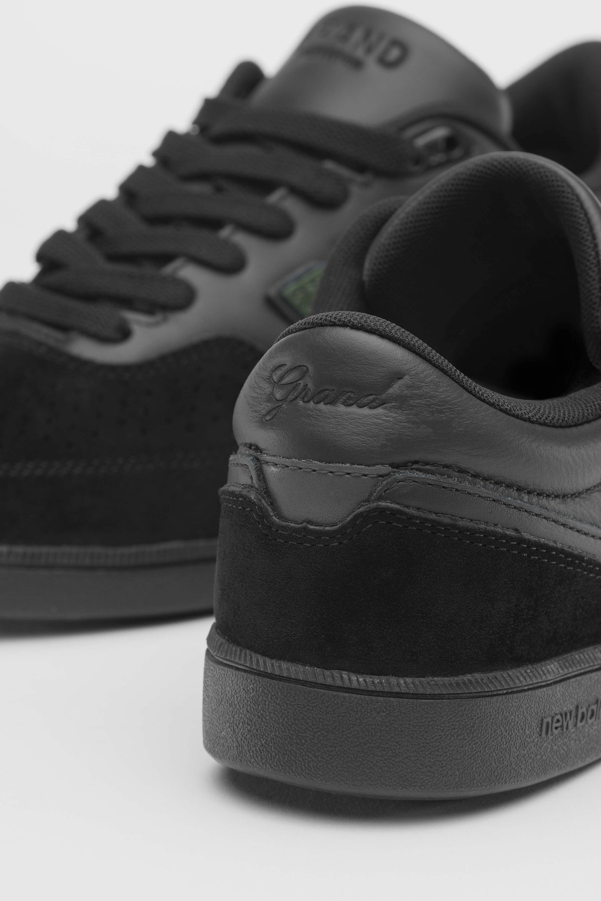 Primer plano del talón de la New Balance Numeric 508 de Grand Collection en negro, con la palabra Grand grabada en letra cursiva sobre el cuero y el logo de New Balance en relieve en la suela