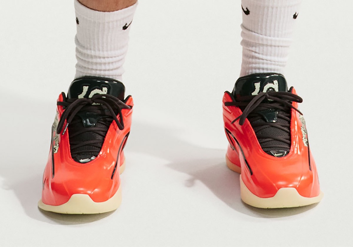 Vista frontal de un par de Nike KD 19 Halloween puestos con medias blancas Nike, con capellada naranja de acabado brillante, cordones marrón oscuro, lengüeta verde oscura con el logo KD en tono claro, un gráfico claro en el lateral y suela beige semitranslúcida, sobre fondo blanco