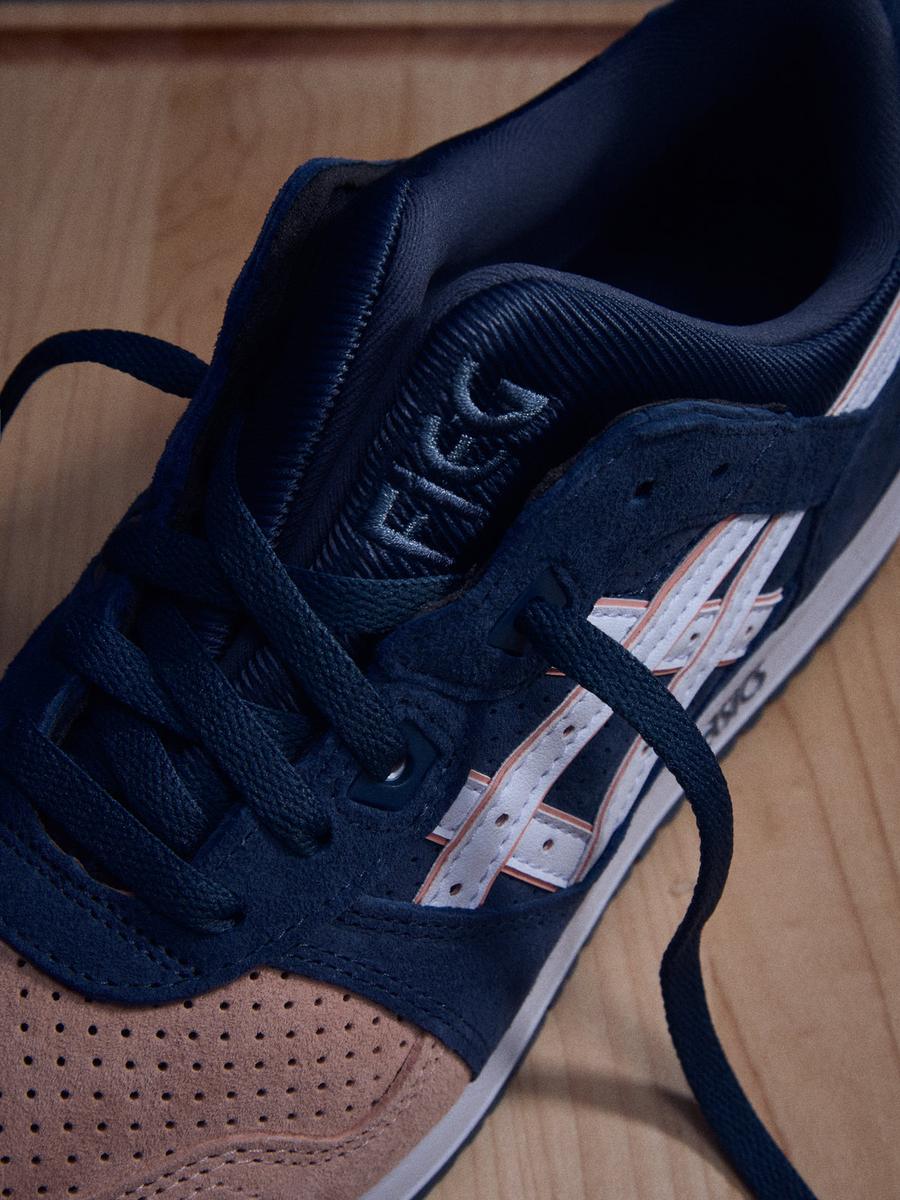 Primer plano de la Kith x ASICS GEL-LYTE III Salmon Toe sobre madera, con la lengüeta partida bordada FIEG y la puntera salmón perforada