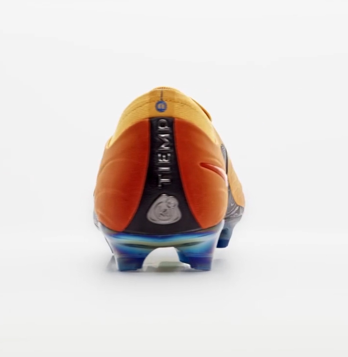 Talón del botín Kids of Immigrants x Nike Tiempo Maestro Elite Sundial sobre fondo blanco, en naranja y rojo, con una franja negra vertical que dice Tiempo y la base de tapones en tonos azules