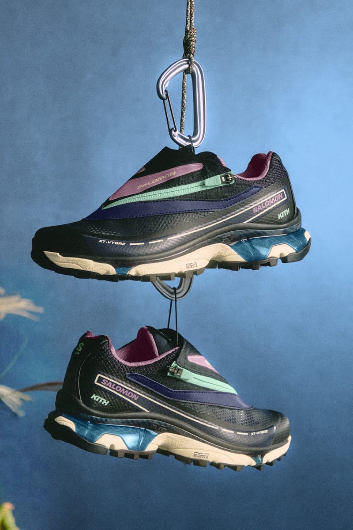 Par de Kith x Salomon XT-VYGR 2 azul marino con detalles lila y verde claro colgando de un mosquetón sobre fondo azul