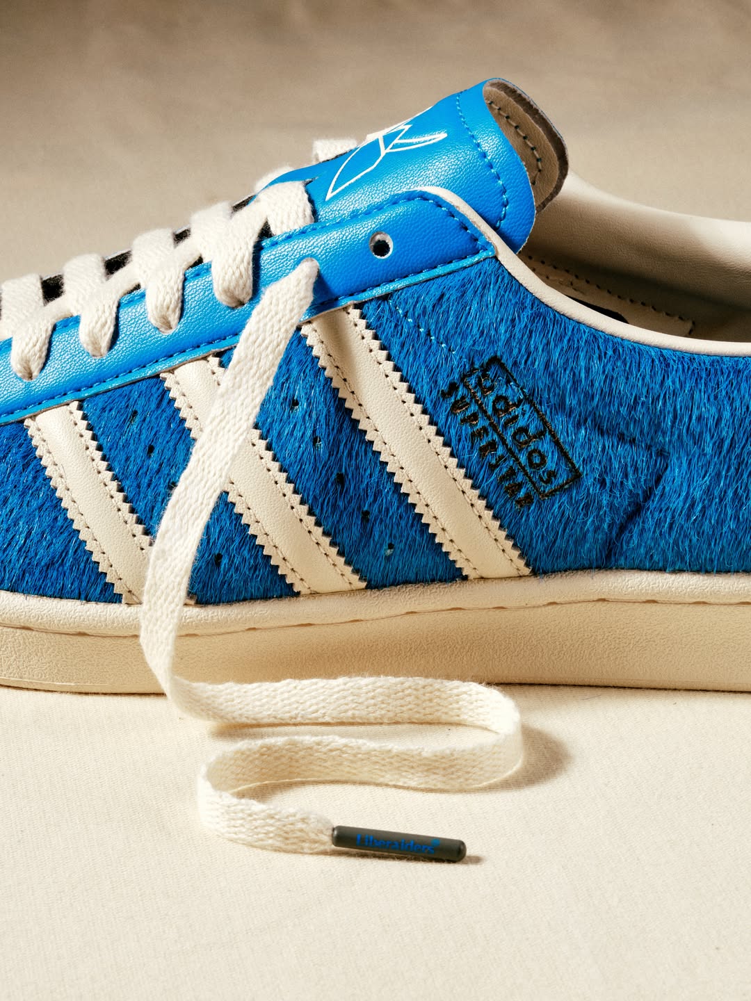 Lateral de la adidas Superstar de Liberaiders con el pelo azul en primer plano, las tres tiras crema, el logo adidas Superstar en el costado y un cordón suelto con el herrete gris que dice Liberaiders