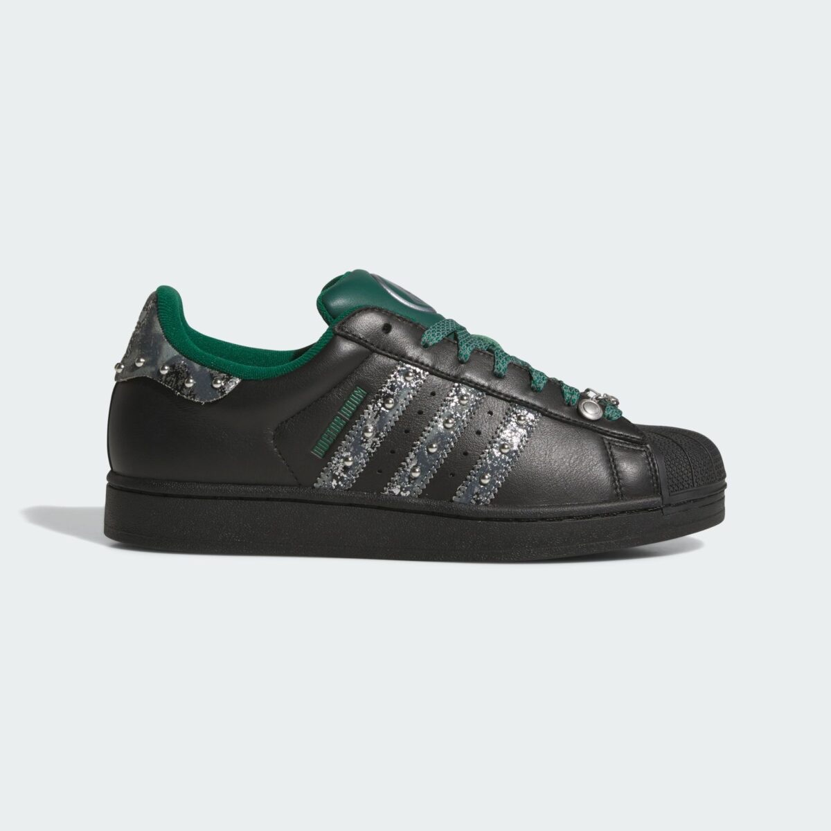 adidas Superstar Doctor Doom de perfil, en cuero negro con tres tiras gris metálico con tachas, cordones y lengüeta verdes y un dije en los ojales