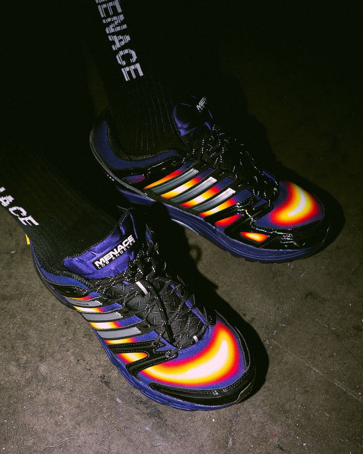 Vista superior de un par de MENACE x adidas Adistar Control 5 Heat Signature puestos con medias negras MENACE, con la puntera encendida en amarillo, rojo y blanco y la base violeta sobre un piso de cemento