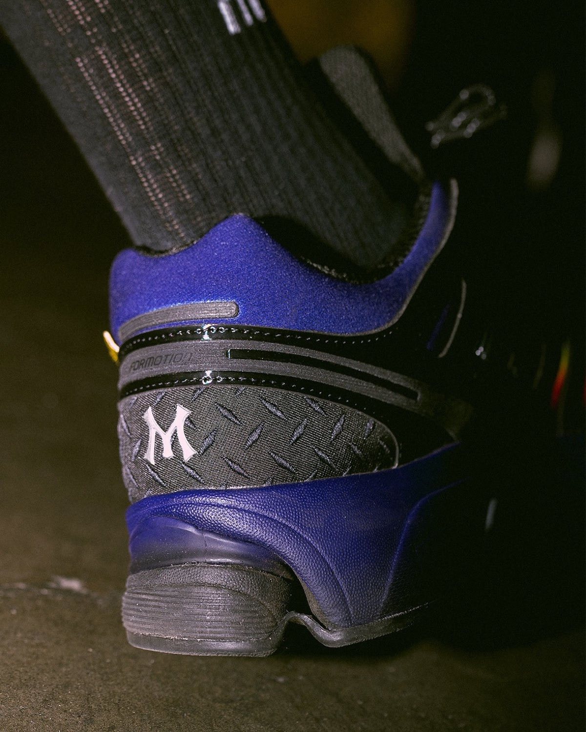 Detalle del talón del MENACE x adidas Adistar Control 5 Heat Signature, con una M blanca sobre un panel negro con textura de chapa antideslizante, cuello violeta y mediasuela violeta