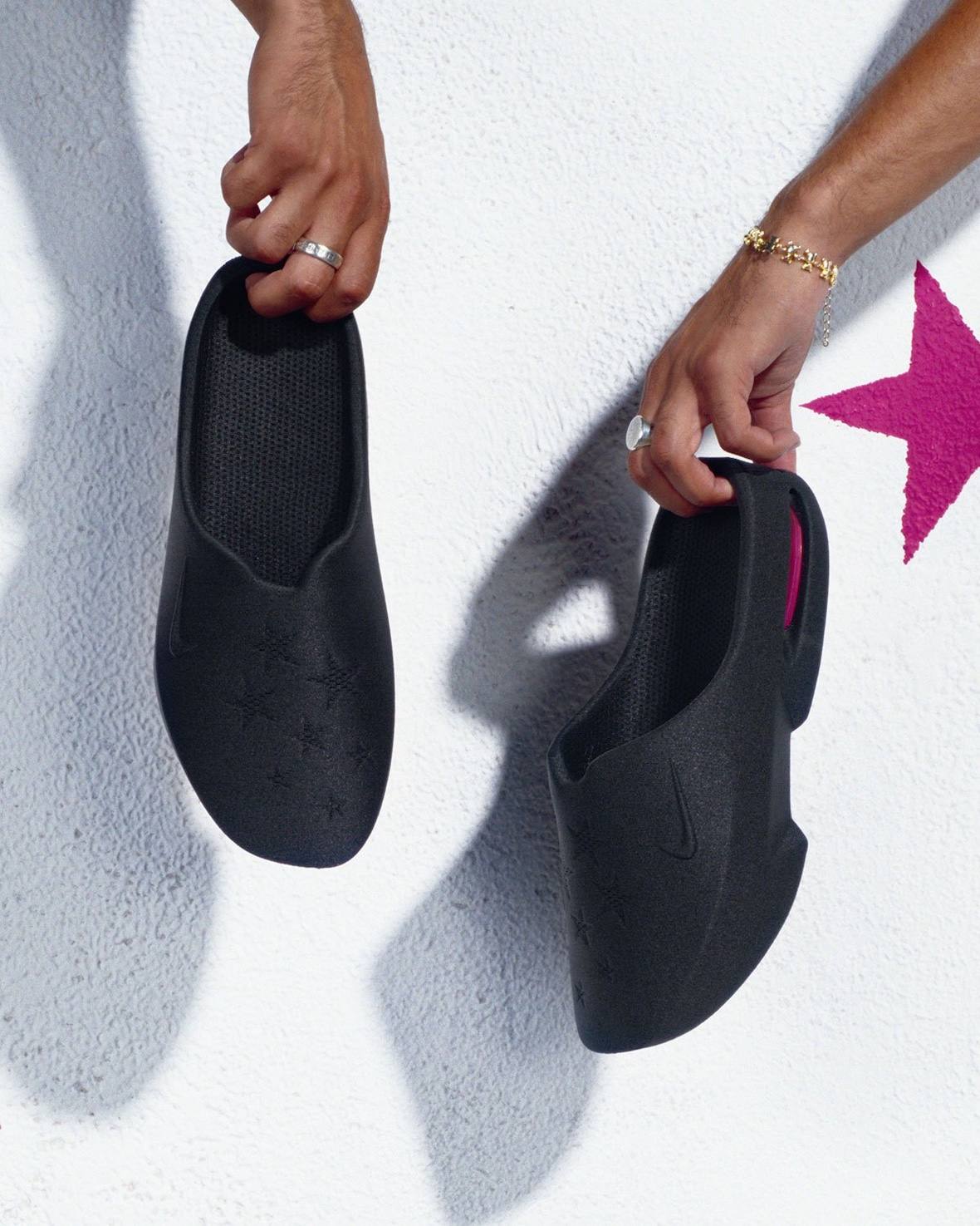 Dos manos sostienen las Nike Air Max Mule negras de Masyn contra una pared blanca, con las estrellas en relieve en la capellada y una estrella rosa pintada en la pared