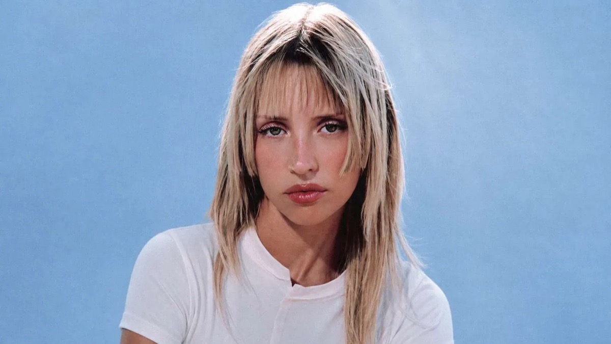 Retrato de prensa de Angèle de frente, con el pelo rubio en capas y flequillo, remera blanca y fondo celeste. Foto: Kenza Le Bas