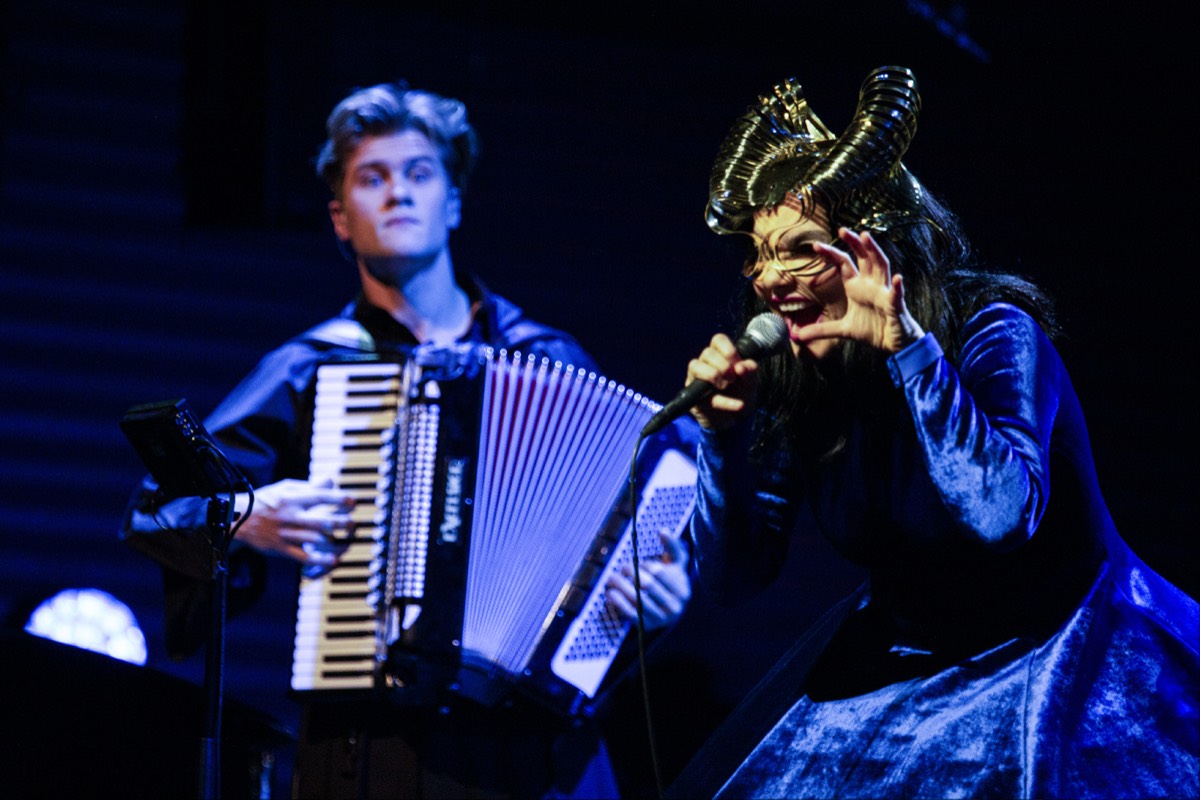 Foto oficial del concierto Orkestral con el coro Hamrahlíð: Björk, con un tocado dorado con forma de cuernos, canta con gesto teatral mientras un músico la acompaña en acordeón