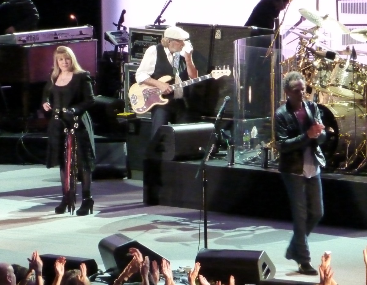 Fleetwood Mac en vivo en el NIA de Birmingham en noviembre de 2009: Stevie Nicks, de negro, a la izquierda; John McVie con el bajo y gorra blanca en el centro, y Lindsey Buckingham, con campera de cuero, caminando hacia el borde del escenario, con el público aplaudiendo abajo (foto de archivo). Foto: Tony Hisgett, CC BY 2.0, vía Wikimedia Commons