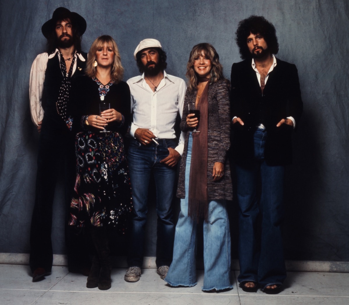 Foto oficial del anuncio de «Fleetwood Mac: The Chain»: Mick Fleetwood, Christine McVie, John McVie, Stevie Nicks y Lindsey Buckingham posan de pie en los años setenta frente a un telón gris azulado, con ropa de época y copas en la mano