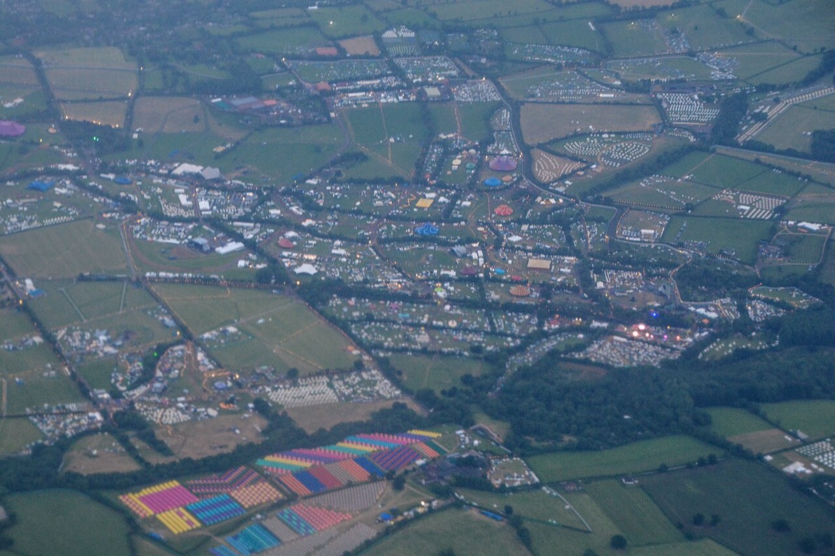 Vista aérea del predio de Glastonbury en Worthy Farm, con campos verdes cubiertos de carpas, carpas de circo de colores y caminos iluminados entre los cercos. Foto: Lewis Clarke, Wikimedia Commons, CC BY-SA 2.0