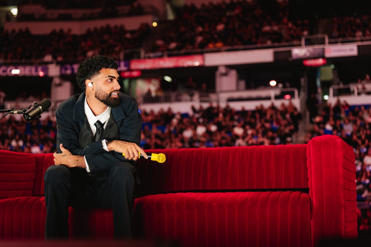 Foto de prensa de Jay Wheeler, de traje oscuro y corbata negra, sentado en un sillón de terciopelo rojo con un micrófono amarillo en la mano, frente a las tribunas llenas de un estadio cubierto