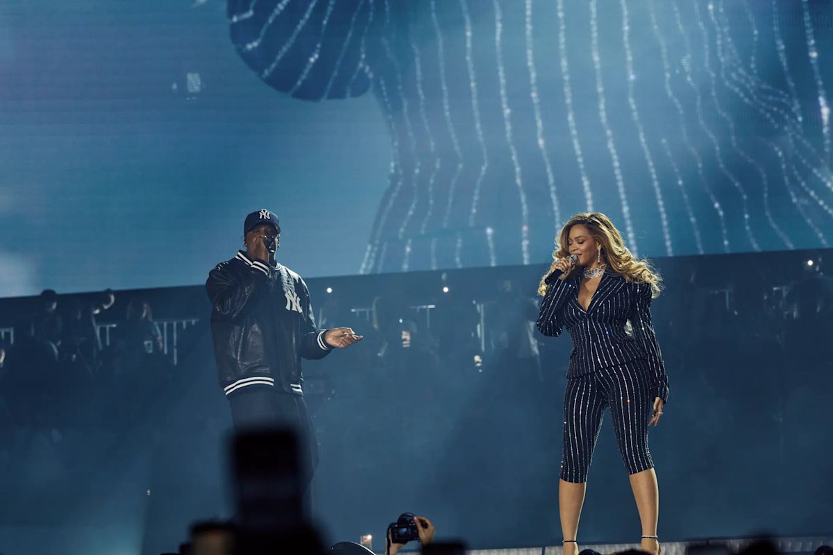 Foto oficial del primer show en el Yankee Stadium: Jay-Z, con gorra y campera de los Yankees, canta junto a Beyoncé, que lleva un traje oscuro a rayas con brillos, frente a una pantalla gigante en tonos azules