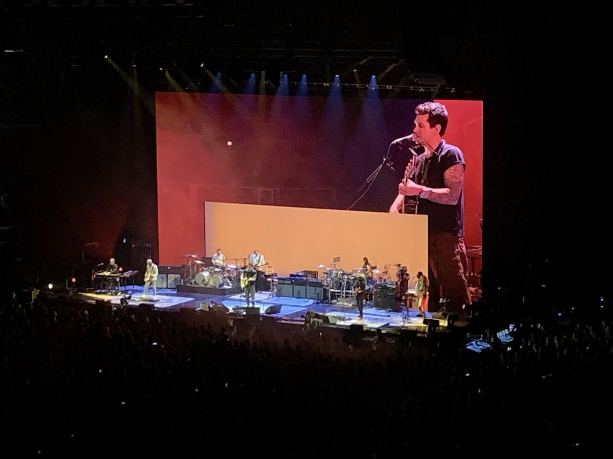 Foto de archivo de John Mayer en un concierto en Chicago, en agosto de 2019: él canta con la guitarra en la punta del escenario, delante de una pantalla roja, mientras la banda toca más atrás. Foto: Cole Camplese, Wikimedia Commons, CC BY 2.0
