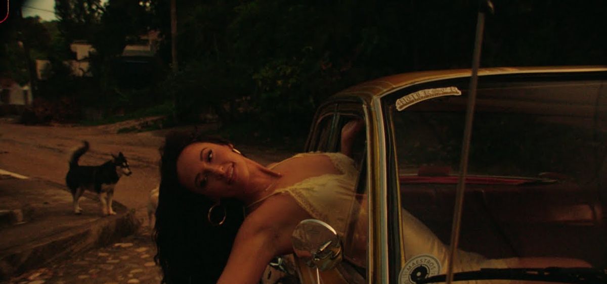 Fotograma de archivo del video oficial de «Mexico Honey», de su disco «Middle of Nowhere»: Kacey Musgraves, con vestido claro, se recuesta contra un auto viejo color dorado en una calle de tierra, con un perro husky al costado