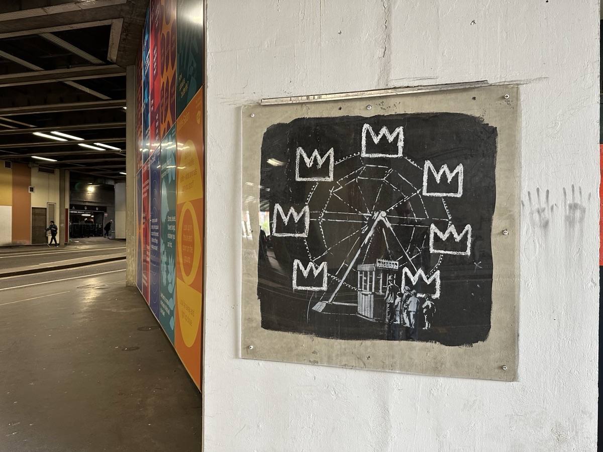 Un mural de Banksy en homenaje a Basquiat: una vuelta al mundo dibujada en blanco con coronas a su alrededor, sobre un fondo negro, protegido por un acrílico en una pared blanca de un pasaje (imagen que representa al artista). Foto: Matt Brown, CC BY 2.0, vía Wikimedia Commons