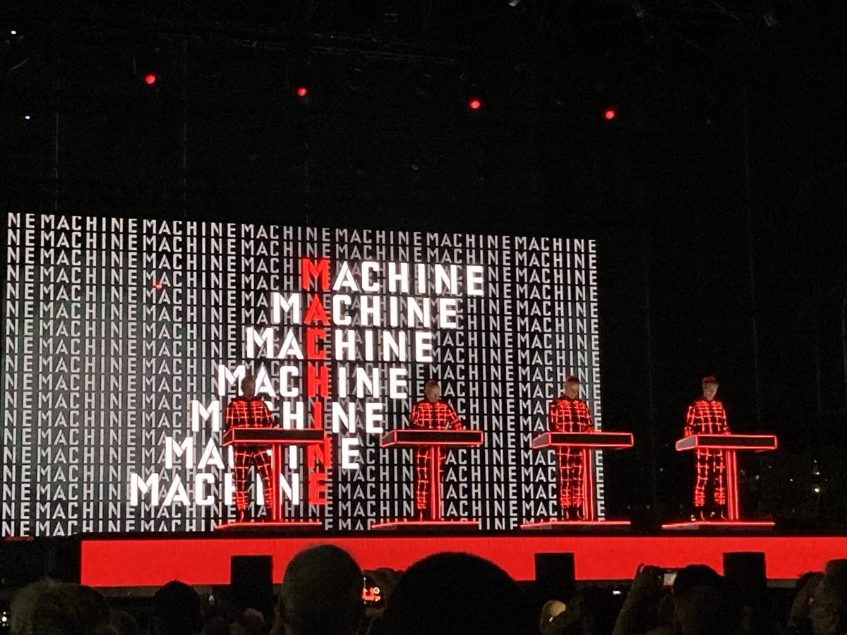 Kraftwerk en vivo en el Arena del Mare de Génova en julio de 2023: los cuatro integrantes con trajes rojos cuadriculados, detrás de sus consolas, frente a una pantalla llena de la palabra MACHINE en blanco y rojo (foto de archivo). Foto: Luca Dell'Orto, CC BY-SA 4.0, vía Wikimedia Commons